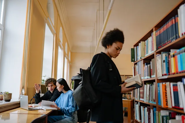 Kitap rafının önünde kitap inceleyen öğrenci ve masada çalışanlar
