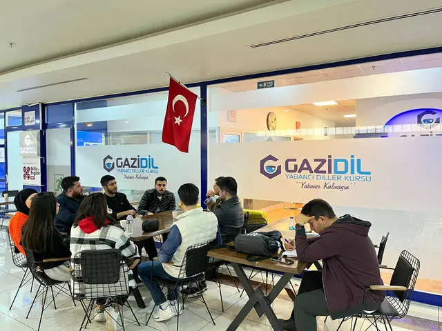 GaziDil logolu cam bölmelerin önünde masalar çevresinde birlikte çalışan katılımcılar