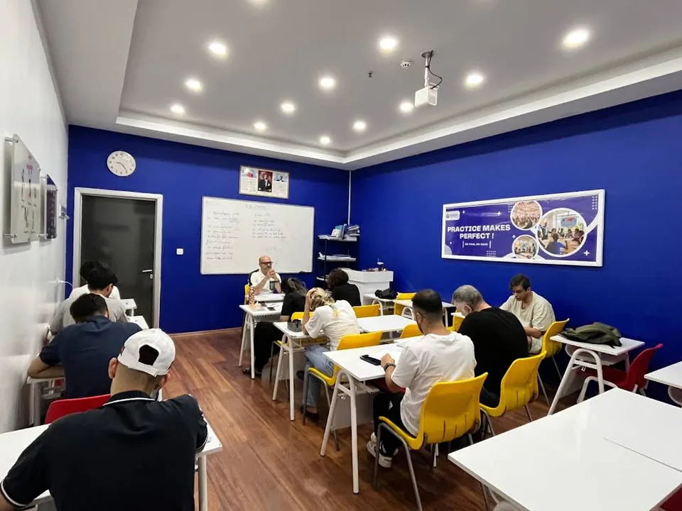 GaziDil’in mavi duvarlı sınıfında beyaz masalar çevresinde derse katılan öğrenciler ve eğitmen